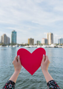Woman holds piece of red paper shaped like a heart in front of St. Petersburg, Florida skyline and ocean