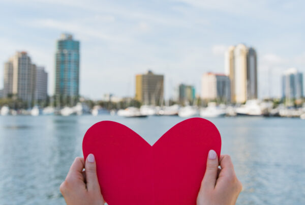 Woman holds piece of red paper shaped like a heart in front of St. Petersburg, Florida skyline and ocean